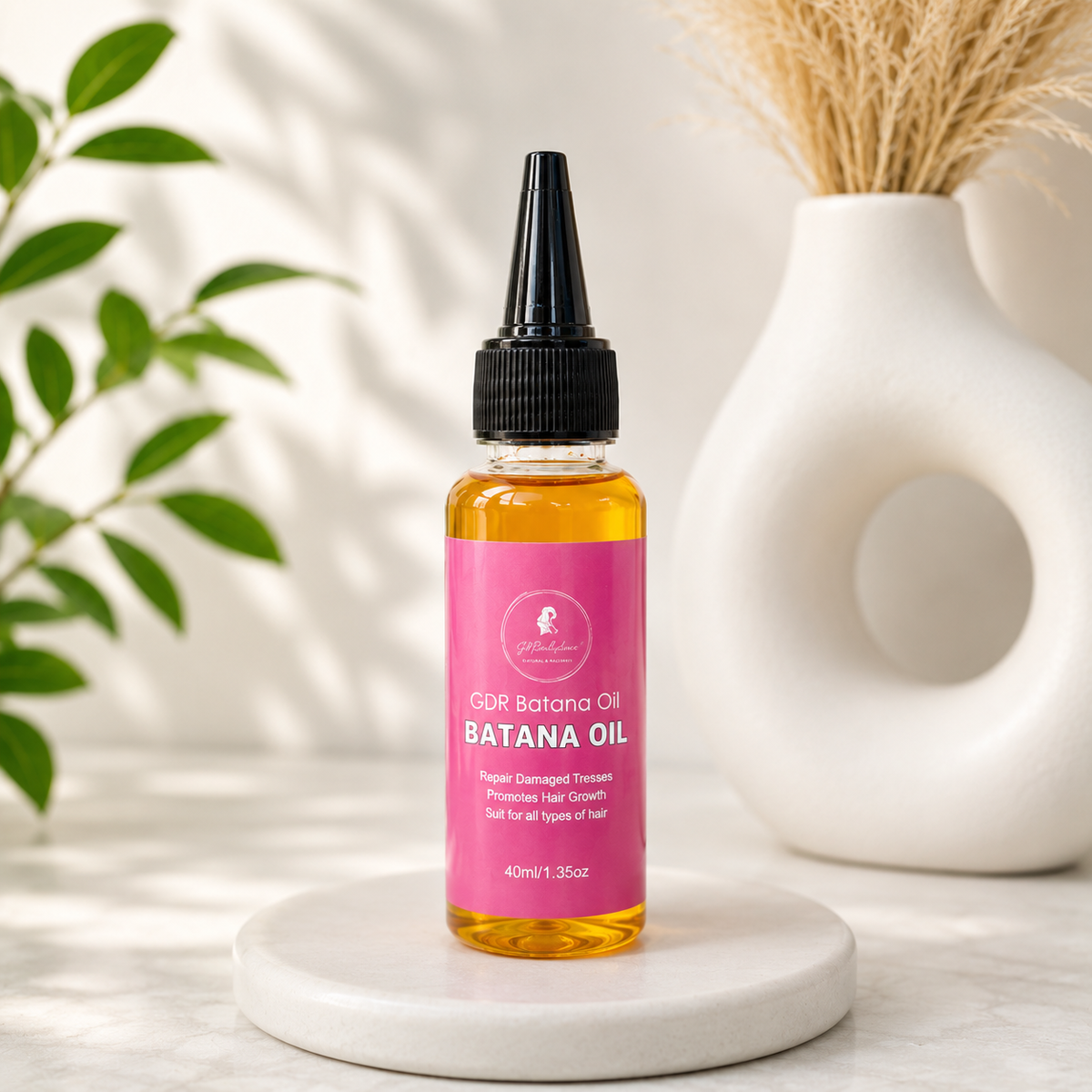 A bottle of GDR Batana Oil with a bright pink label and black dropper cap, displayed on a white marble coaster with a white ceramic vase and green leaves in the background.
