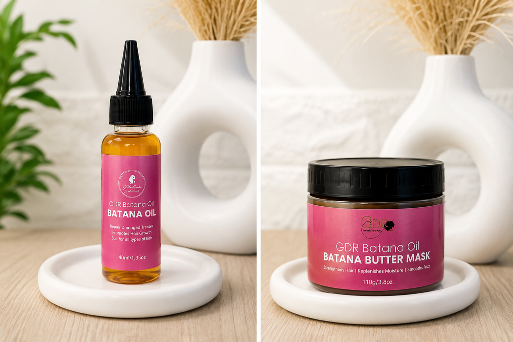 Two GDR haircare products displayed side by side on white ceramic stands: a small bottle of Batana Oil with amber liquid and a pink label, and a jar of Batana Butter Mask with a black lid and matching pink label. The background shows a light wood surface, white decorative vases with dried grass, and a blurred plant.