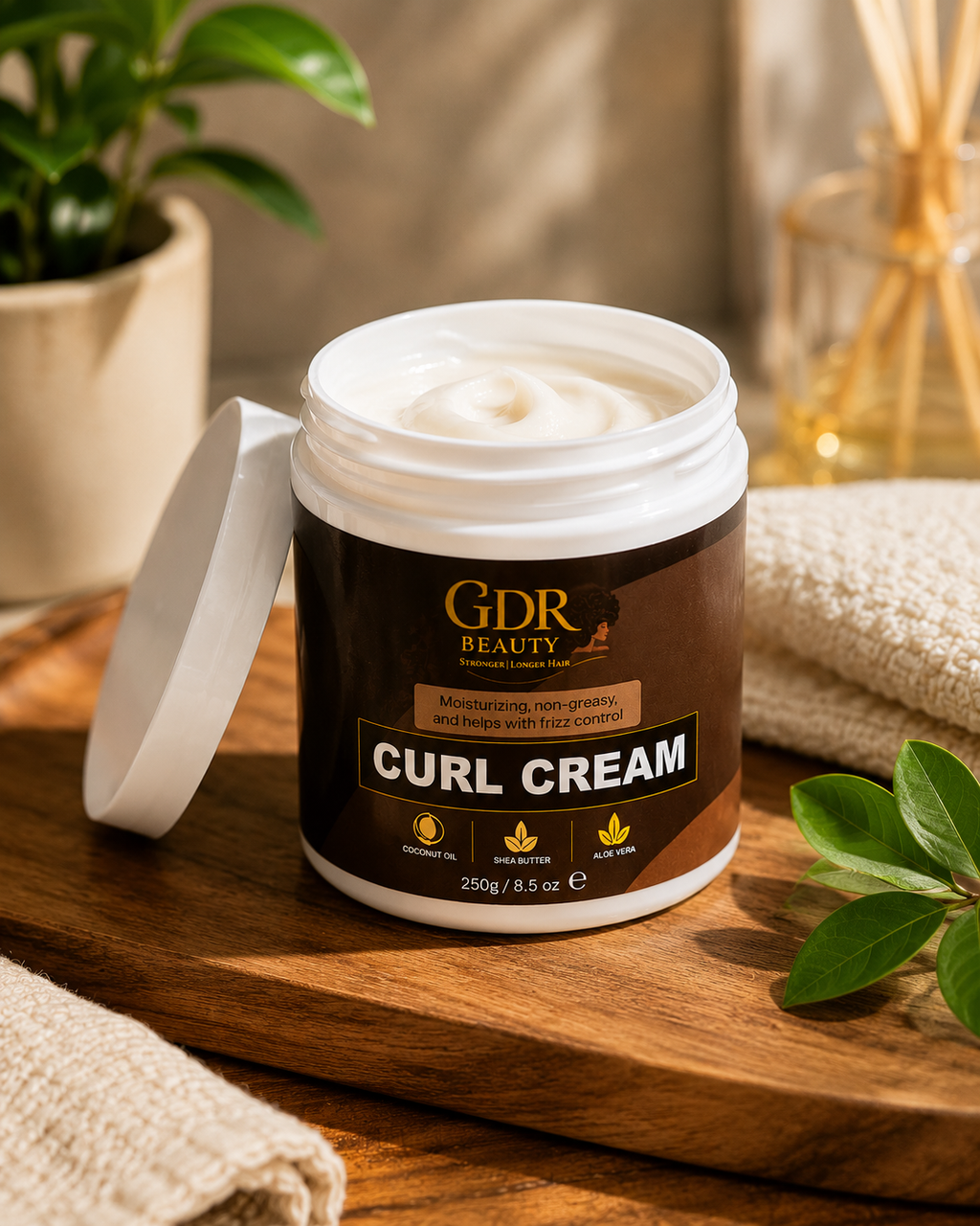 An open jar of GDR Beauty Curl Cream with white cream visible inside, set on a wooden surface with a lid leaning beside it. The scene includes a potted plant, rolled towels, and green leaves in a styled, warm environment.