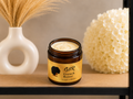An open amber glass jar of GDR Growth Butter with a yellow label, displayed on a wooden shelf next to a white vase with dried grass and a white flower ball.