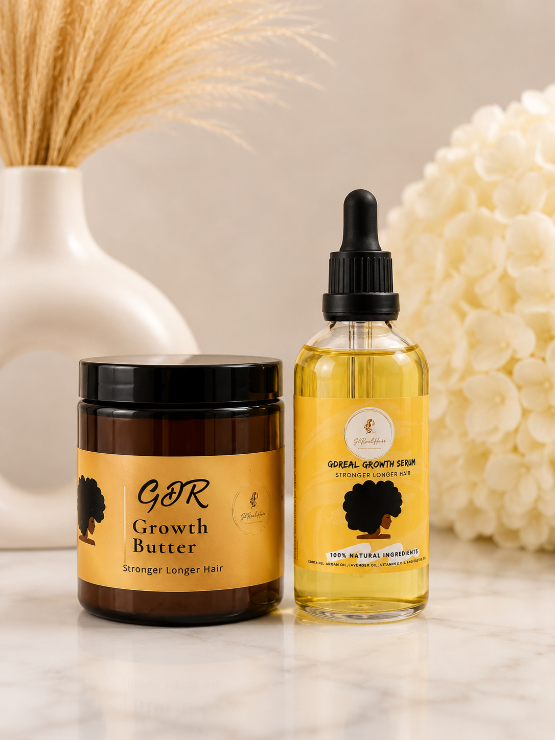 A jar of GDR Growth Butter and a bottle of GDR Growth Serum displayed side-by-side on a marble surface with dried wheat and white flowers in the background.