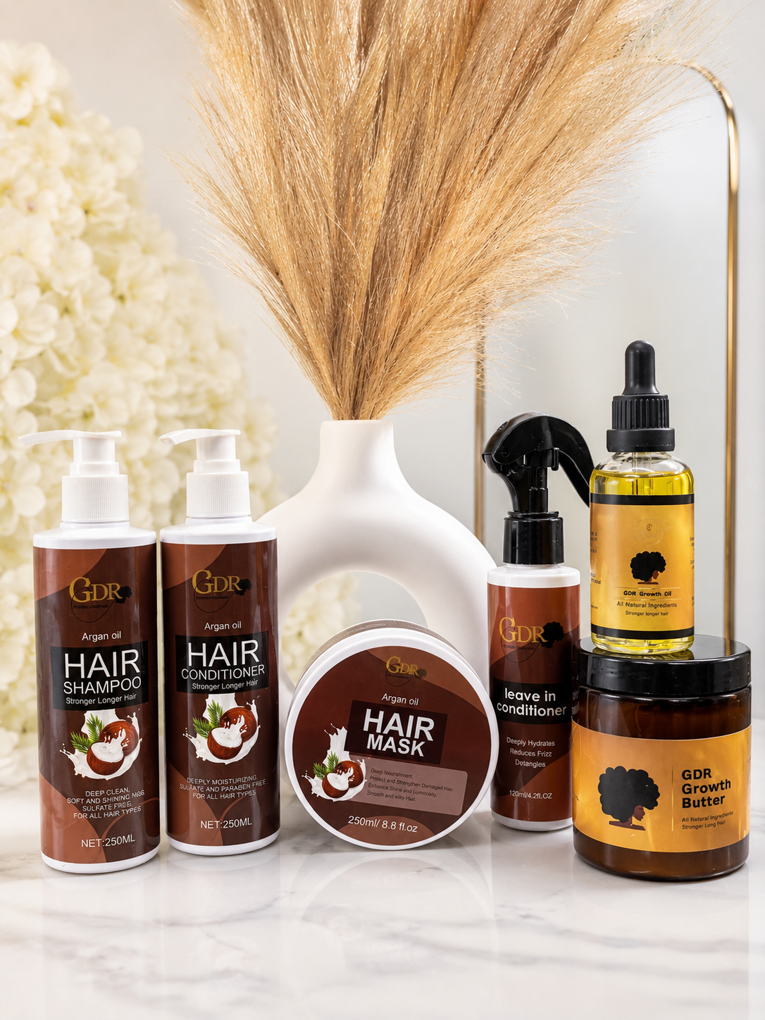 A GDR hair care set arranged on a white marble surface with a white vase of dried grass behind it. The products include two pump bottles of Hair Shampoo and Hair Conditioner, a jar of Hair Mask, a spray bottle of Leave-In Conditioner, and a jar of GDR Growth Booster.