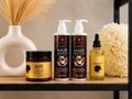 A set of hair care products arranged on a wooden shelf: two pump bottles of Argan oil Hair Shampoo and Hair Conditioner, a jar of GDR Growth Butter, and a dropper bottle of Argan Oil Hair Serum. Decorative dried grass in a white vase and white flowers sit beside the products.