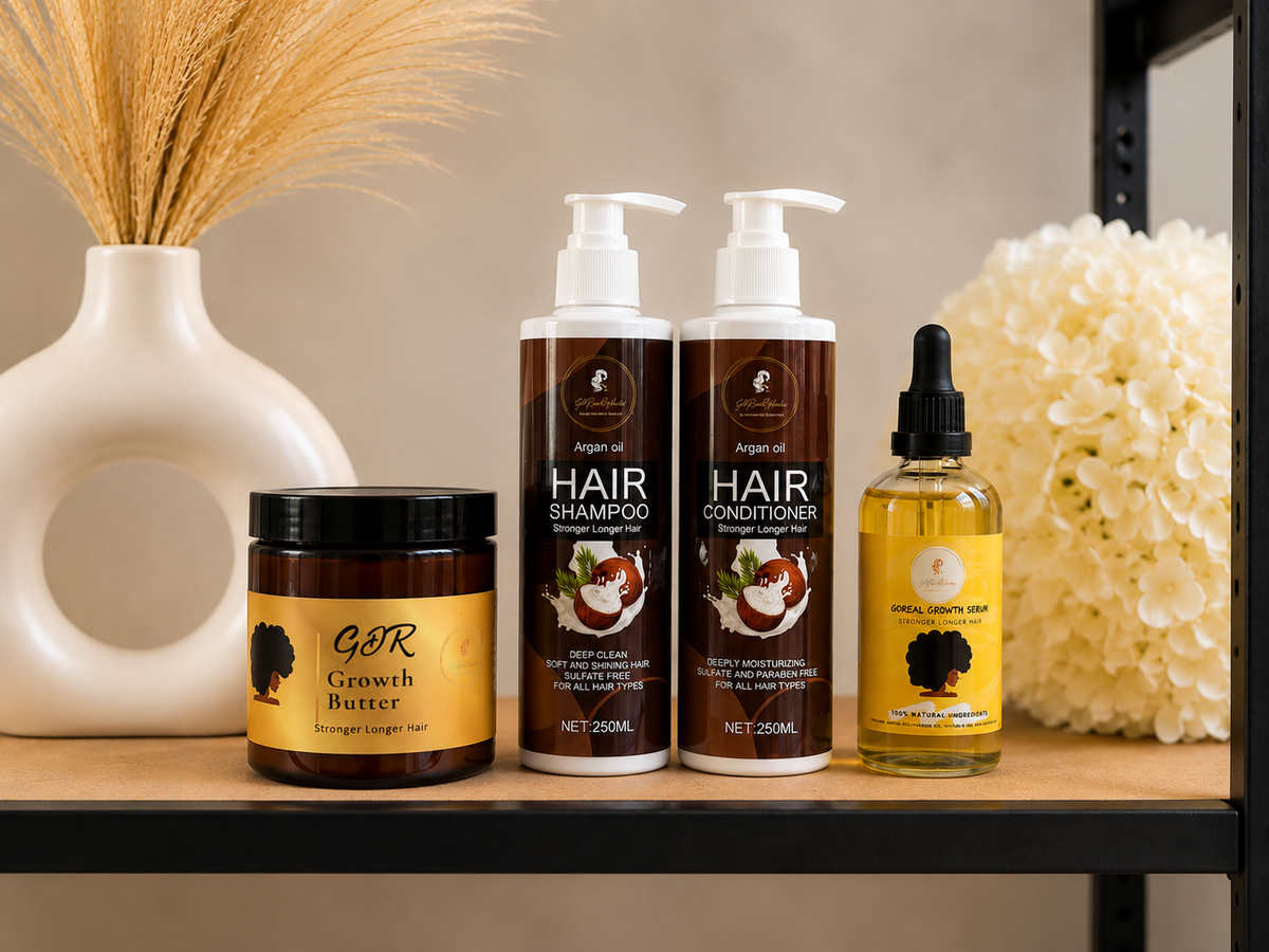 A set of hair care products arranged on a wooden shelf: two pump bottles of Argan oil Hair Shampoo and Hair Conditioner, a jar of GDR Growth Butter, and a dropper bottle of Argan Oil Hair Serum. Decorative dried grass in a white vase and white flowers sit beside the products.
