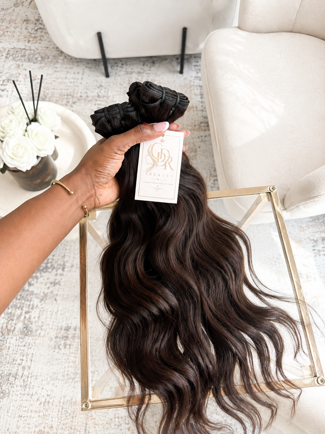 A hand holds a bundle of long, dark brown body wave hair extensions with a white brand tag, displayed over a gold-framed glass tray in a bright, modern room setting.