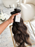 A hand holds a bundle of long, dark brown wavy hair extensions with a white brand tag, set against a bright, modern interior background with a cream chair and a gold-framed tray.