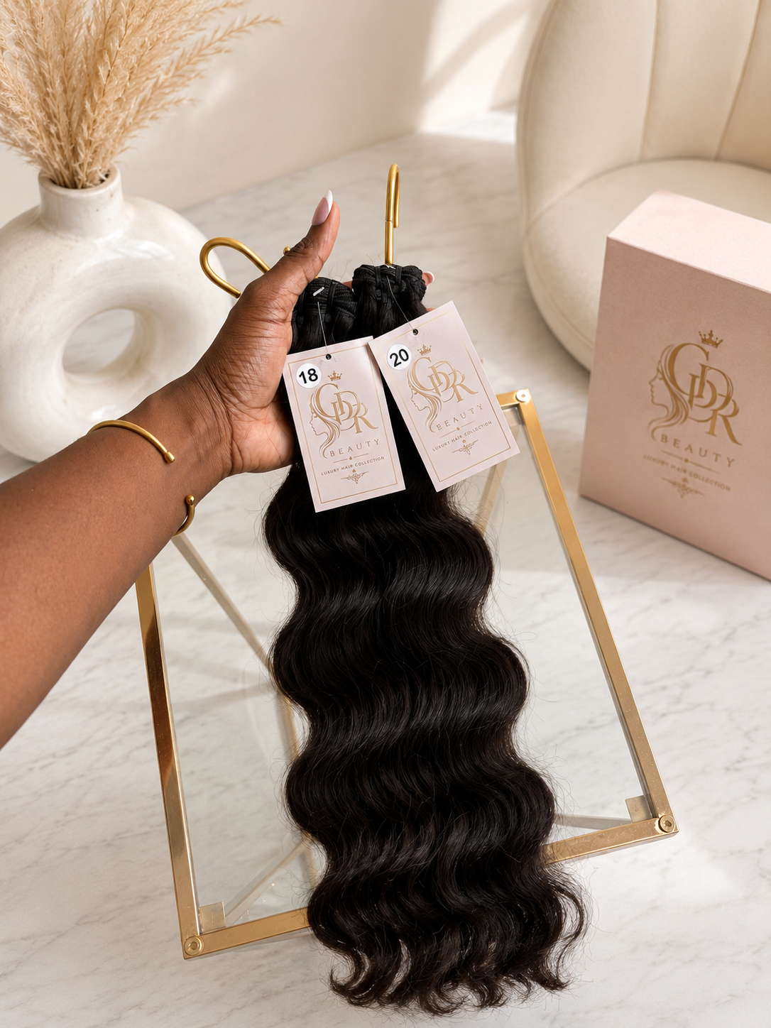 A hand holds a bundle of long, dark brown to black body wave hair extensions with tags, displayed on a gold stand next to a pink hair box with a gold logo.